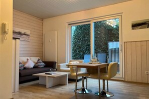 A modern living room with a sofa, coffee table, dining table, and chairs. There is a large window providing a view of greenery outside. - Holiday apartment in Föhr with 1 bedroom (Wyk auf Föhr)