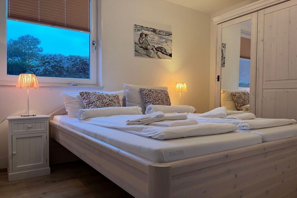 Een opgemaakt bed met witte lakens en handdoeken, een nachtkastje met een lamp, een raam met zonwering en een schilderij aan de muur.