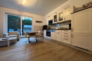 A modern kitchen with white cabinetry, a dining area with a table and chairs, and a large sliding glass door leading to a garden. - Holiday apartment in Föhr with 1 bedroom (Wyk auf Föhr)