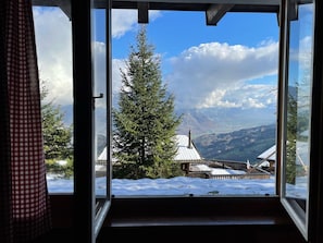 A view through a window with snow-covered ground, a large evergreen tree, and a building with a sloped roof. - Chalet Felsenberg (CharmingStay), (Flumserberg Tannenheim), by Interhome (Flumserberg Tannenheim)