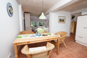 A dining area with a wooden table, wicker chairs, a hanging light fixture, and a kitchen in the background. - Holiday home in Borgsum with swimming pool (Borgsum)