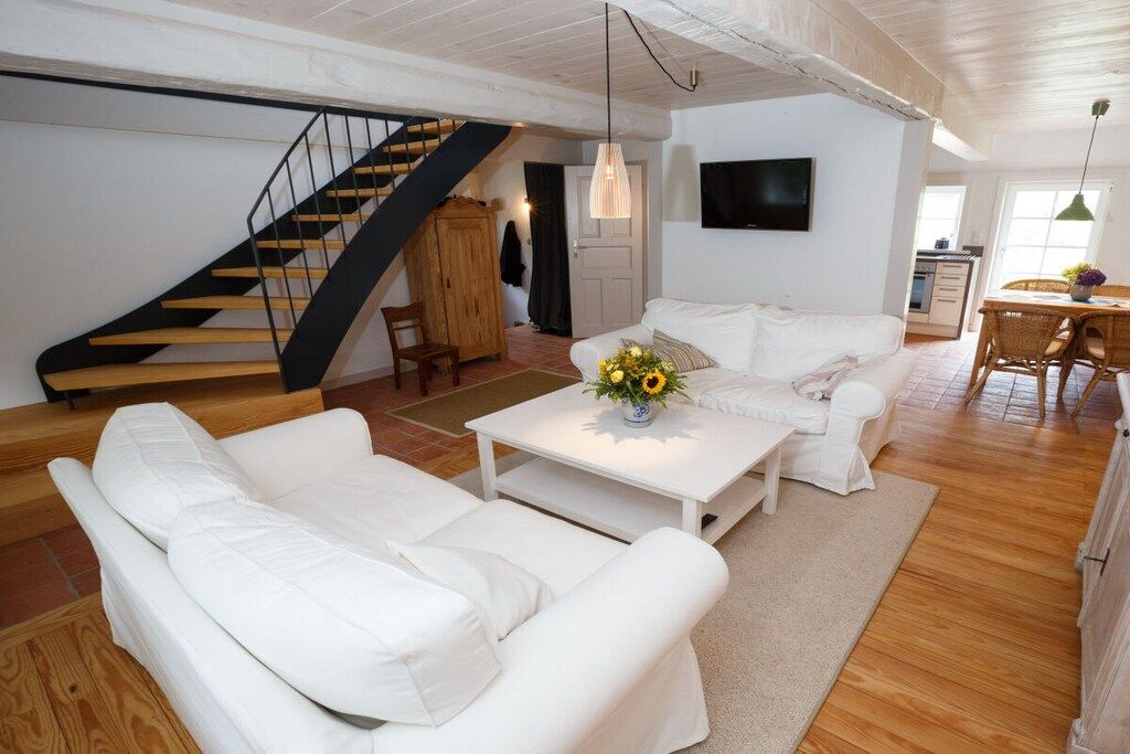 A spacious living area with a white sofa set, a coffee table with a floral centerpiece, a wooden staircase, and a kitchen in the background.
