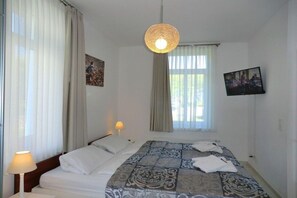 A hotel room with a bed, a hanging light fixture, a window with curtains, and a television on the wall. - Holiday apartment with 1 bedroom (Wyk auf Föhr)