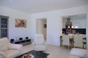 A modern living room with a sofa, armchair, and coffee table. There is a dining area with chairs and a table. A kitchen is visible in the background. - Holiday apartment with 1 bedroom (Wyk auf Föhr)