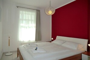A hotel room with a bed, a bedside table, a lamp, a window with curtains, and a red accent wall. - Holiday apartment with 1 bedroom (Wyk auf Föhr)