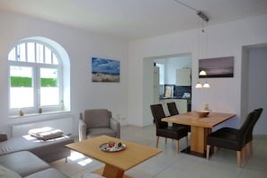 A modern living room with a sofa, a dining table with chairs, a large window, and artwork on the walls. - Holiday apartment with 1 bedroom (Wyk auf Föhr)