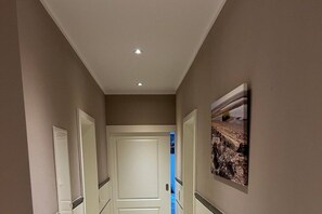 Un couloir avec une porte blanche au fond, un tableau encadré au mur et un luminaire au plafond.