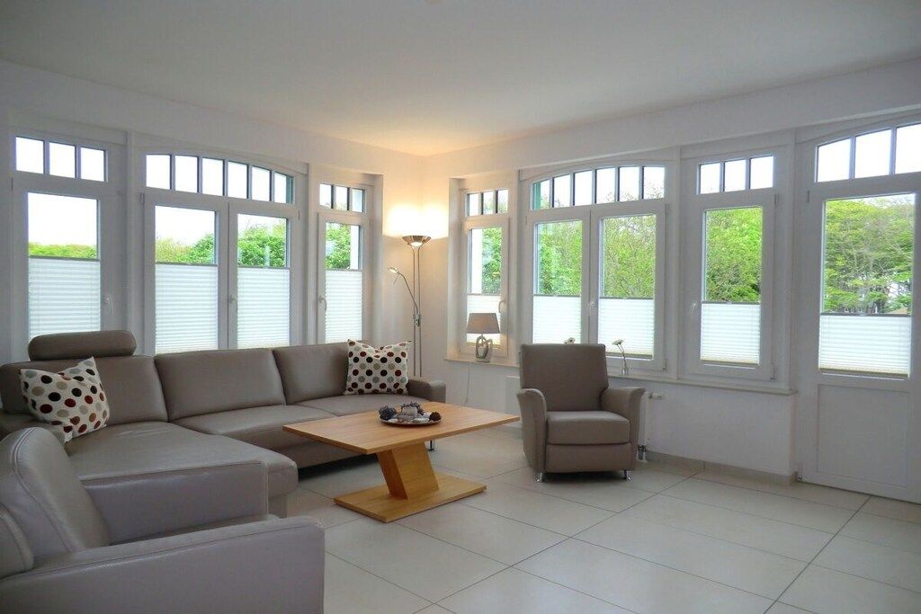Een moderne woonkamer met een hoekbank, een salontafel en een fauteuil. Grote ramen met jaloezieën, een vloerlamp en uitzicht op groen buiten.