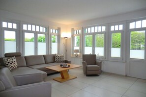 A modern living room with a sectional sofa, a coffee table, and a chair. There are large windows with blinds, a floor lamp, and a view of greenery outside. - Holiday apartment with 1 bedroom (Wyk auf Föhr)