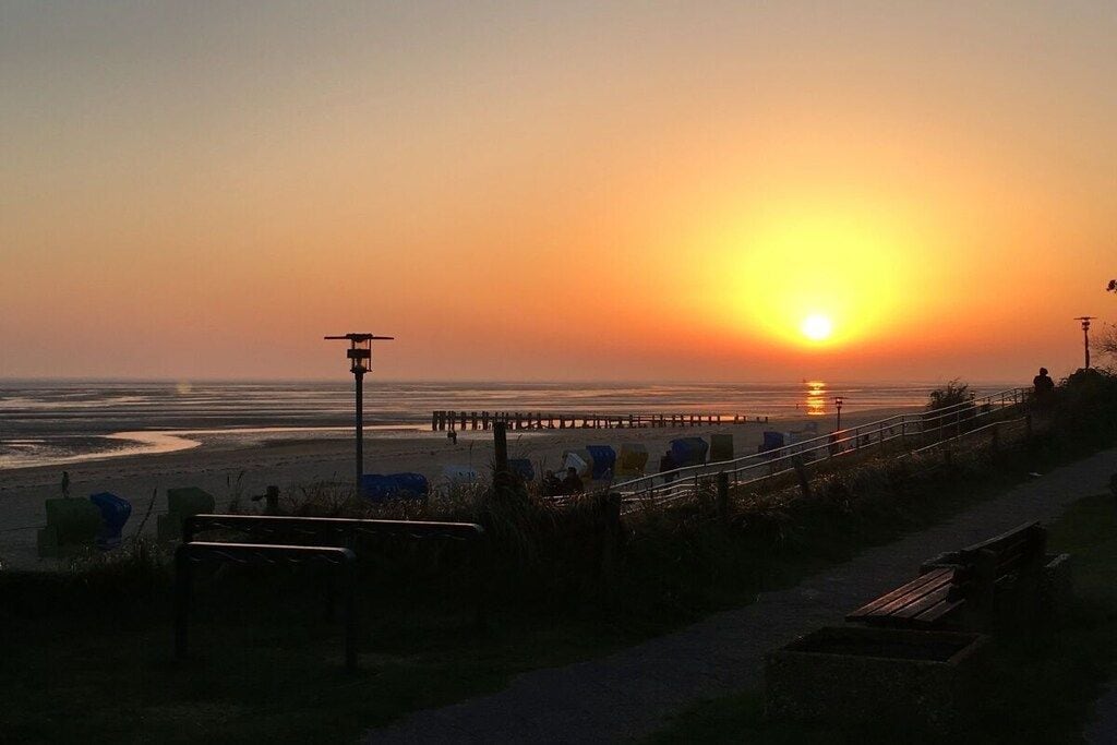 A sunset over a beach with a pier extending into the water, a walkway, and benches.