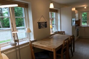 A dining area with a wooden table and chairs, a window with a view of greenery, and a wall-mounted clock. - Green Eilun - House Section 5 (Wyk auf Föhr)