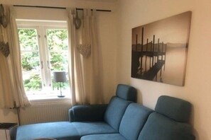 A room with a blue sofa, a window with curtains, a radiator, and a painting on the wall. - Green Eilun - House Section 5 (Wyk auf Föhr)