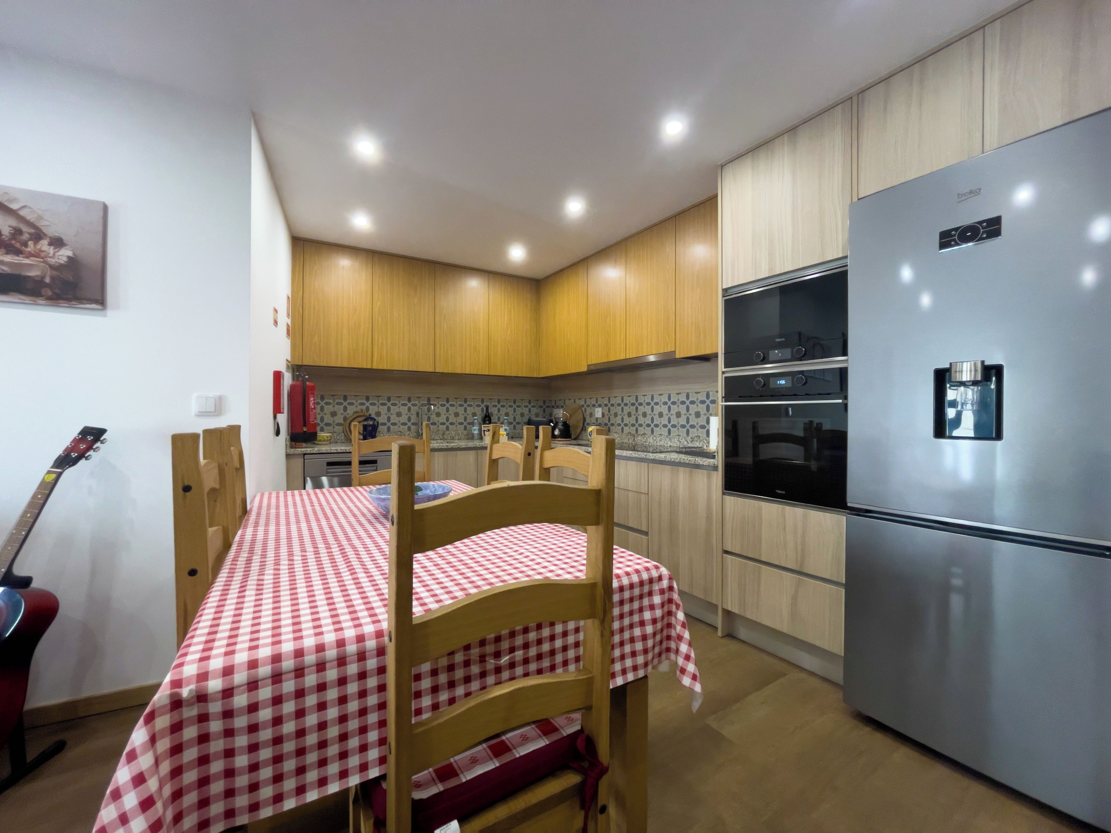 A kitchen with wooden cabinets, a stainless steel refrigerator, a checkered tablecloth, and a guitar leaning against the wall.