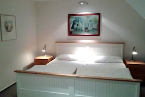 Une chambre à coucher comprenant un lit, deux tables de chevet, une commode, un tableau encadré au mur et un luminaire au plafond.