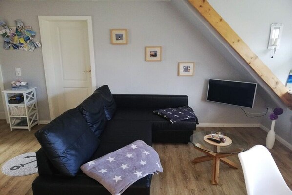 Ein Wohnzimmer mit einem schwarzen Sofa, einem Glastisch, einem weißen Sessel und einem Wand-Fernseher.