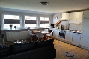 Eine kleine Küche mit weißen Schränken, ein Essbereich mit rundem Tisch und Stühlen sowie ein schwarzes Sofa.
