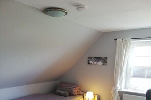 A bedroom with a sloped ceiling, a bed with a patterned bedspread, a bedside lamp, a window with curtains, and a wall-mounted picture. - Holiday apartment in Föhr with 1 bedroom (Nieblum)