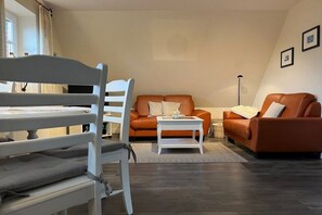 A living room with a white chair, an orange sofa, a white coffee table, and a floor lamp. - Holiday apartment in Föhr with 1 bedroom (Nieblum)