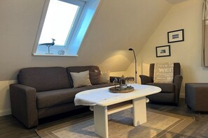 A cozy living room with a sofa, a coffee table, and a lamp. There is a window with a view of the sky and a small bird figurine on the sill. - Holiday apartment in Föhr with 1 bedroom (Nieblum)