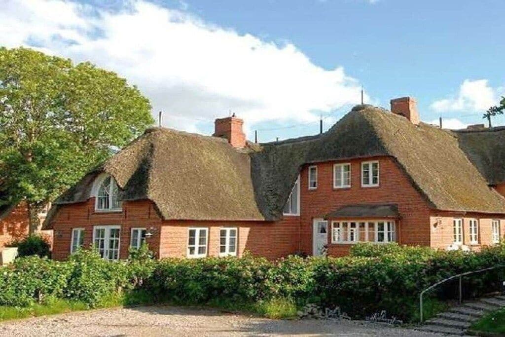 A traditional thatched-roof house with a brick exterior, a visible chimney, and a gravel driveway leading to a set of steps.