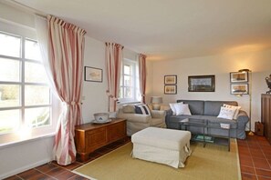Una sala de estar con sofá, reposapiés y una mesa de centro. Hay cortinas en las ventanas y una lámpara en la pared.
