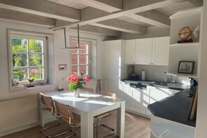 A modern kitchen with white cabinetry, a dining table with chairs, and a window with a view of greenery. - Holiday home in Oldsum with swimming pool (Oldsum)