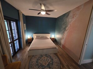 Ein Schlafzimmer mit einem Bett, zwei Nachttischlampen, einem Deckenventilator, einem großen Fenster mit Vorhängen und einer strukturierten Wand.