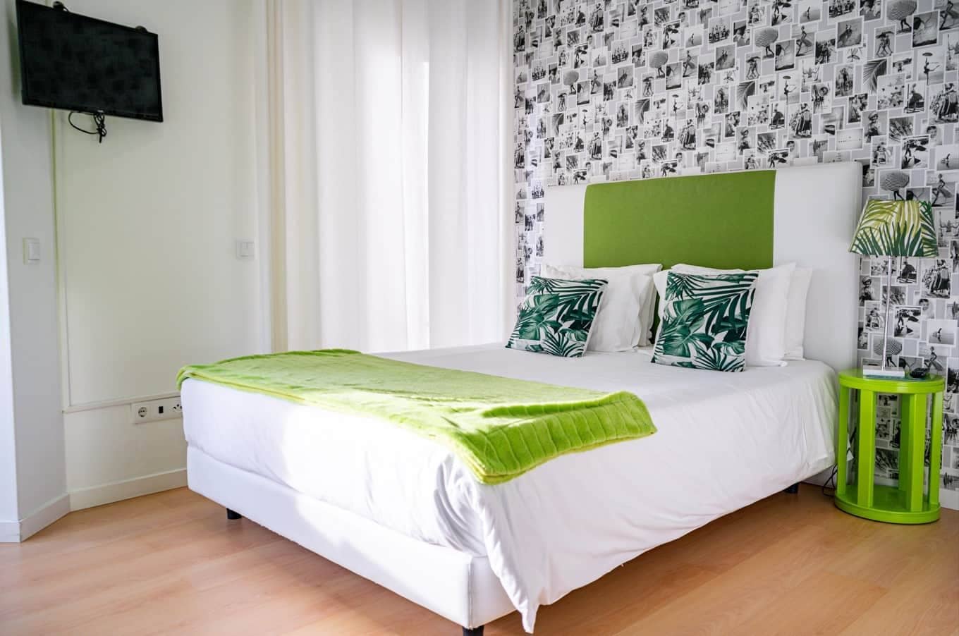 Un dormitorio con una cama, un cabecero verde, ropa de cama blanca, una manta verde y una pared con un collage de fotografías blanco y negro.