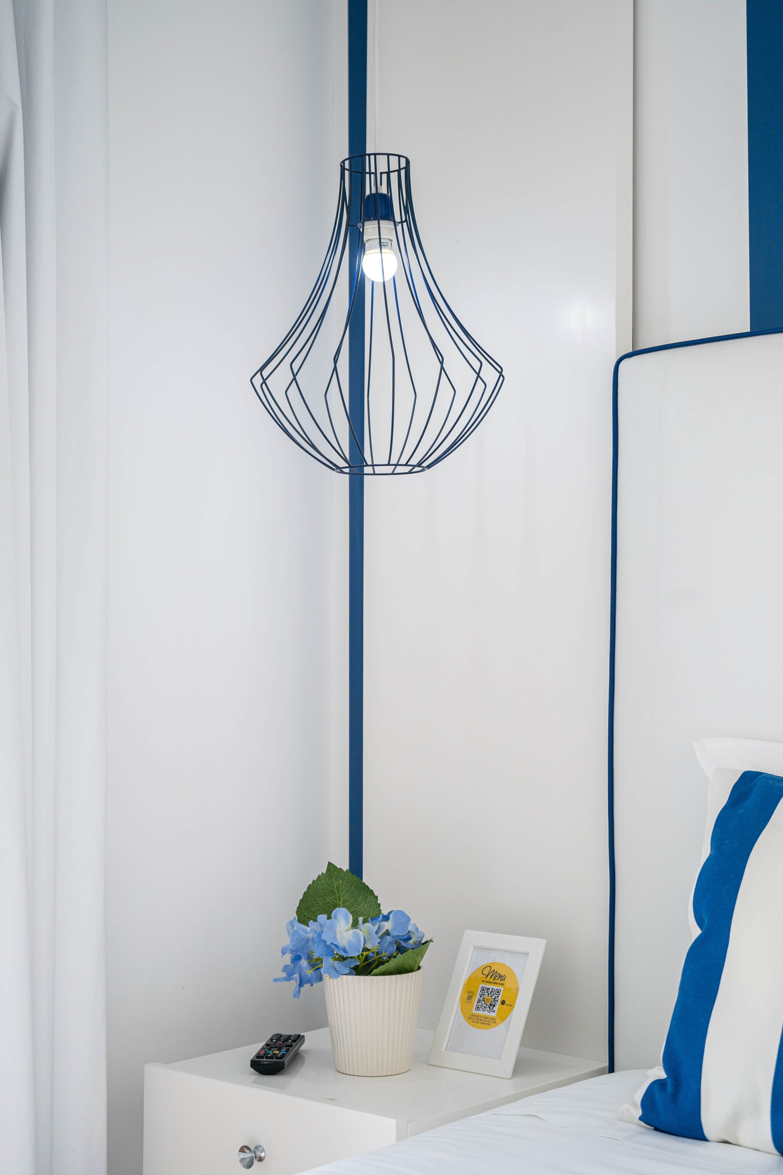 A modern bedroom with a white bed, a blue and white pillow, a white nightstand, a remote control, a framed picture, a potted plant, and a unique black wireframe lamp.