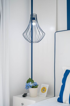 Une chambre moderne avec un lit blanc, un oreiller bleu et blanc, une table de chevet blanche, une télécommande, un tableau encadré, une plante en pot et une lampe originale à structure en fil de fer noir.