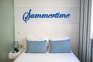 A bed with two pillows, a wall-mounted sign reading 'Summertime,' a bedside table with a remote control, and a striped curtain. - Double Room Street View (Nazaré)