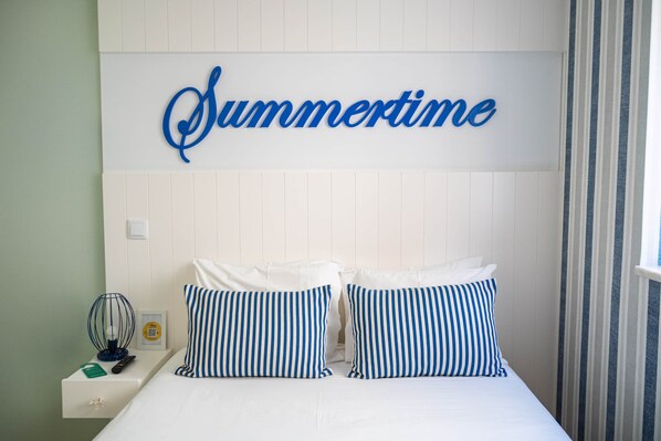 Una cama con dos almohadas, un cartel en la pared que dice 'Summertime', una mesita de noche con un control remoto y una cortina a rayas.