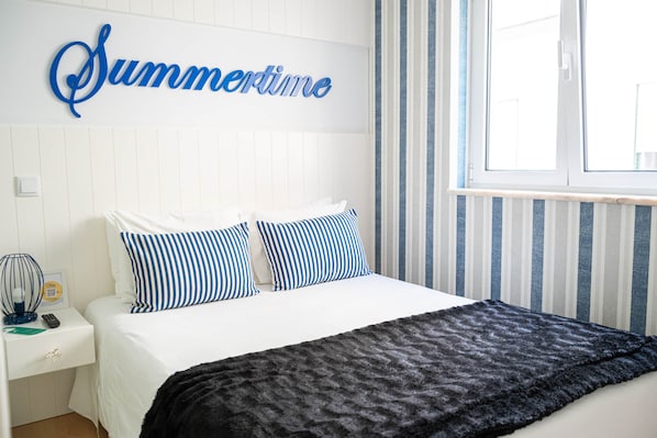 A bedroom with a bed, pillows, a bedside table, a lamp, and a window with curtains. - Double Room Street View (Nazaré)