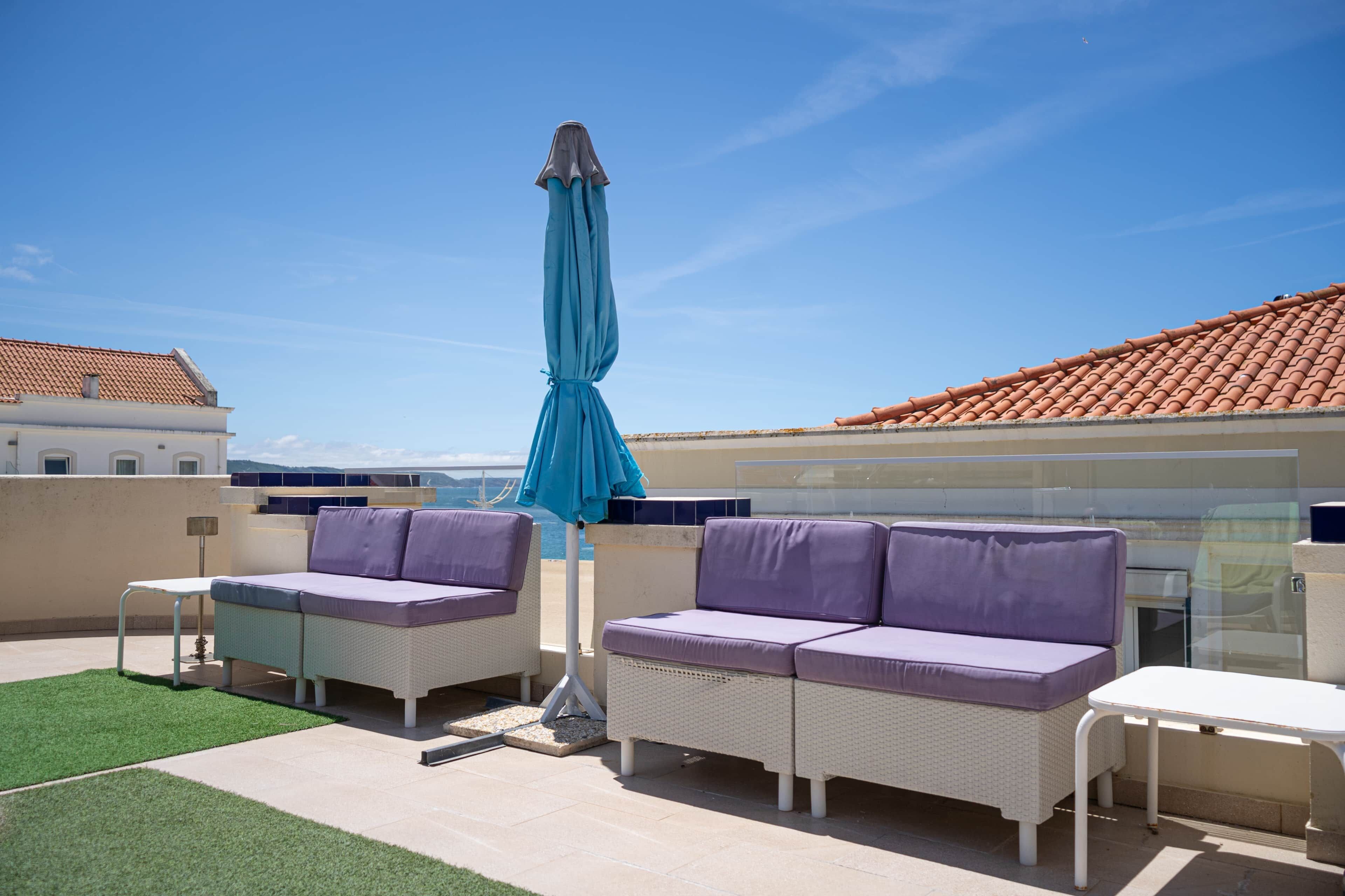An outdoor seating area with two sofas and a table, a blue umbrella, and a view of the sea.