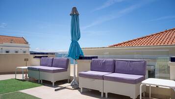 An outdoor seating area with two sofas and a table, a blue umbrella, and a view of the sea.