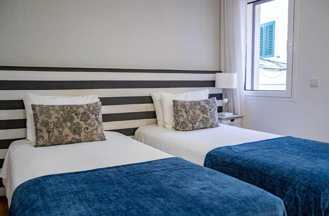 Een hotelkamer met twee bedden, een gestreept hoofdeinde, een nachtkastje met een lamp en een raam met uitzicht op gebouwen.