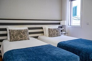 A hotel room with two beds, a striped headboard, a bedside table with a lamp, and a window with a view of buildings. - Double Triple Suite with Balcony and Side Ocean View (Nazaré)