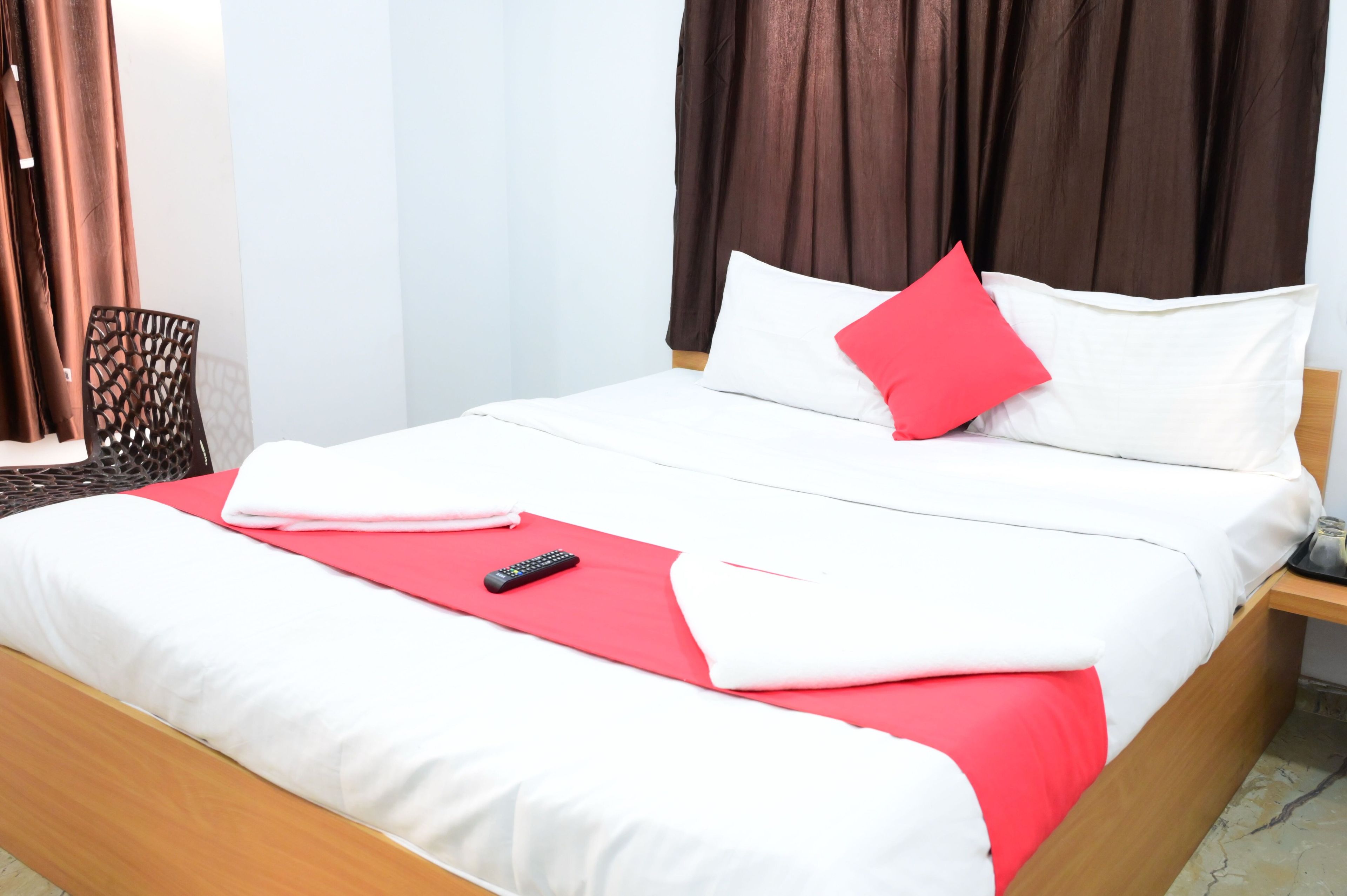 Una cama bien hecha con ropa de cama blanca y roja, un control remoto y una silla con respaldo estampado.