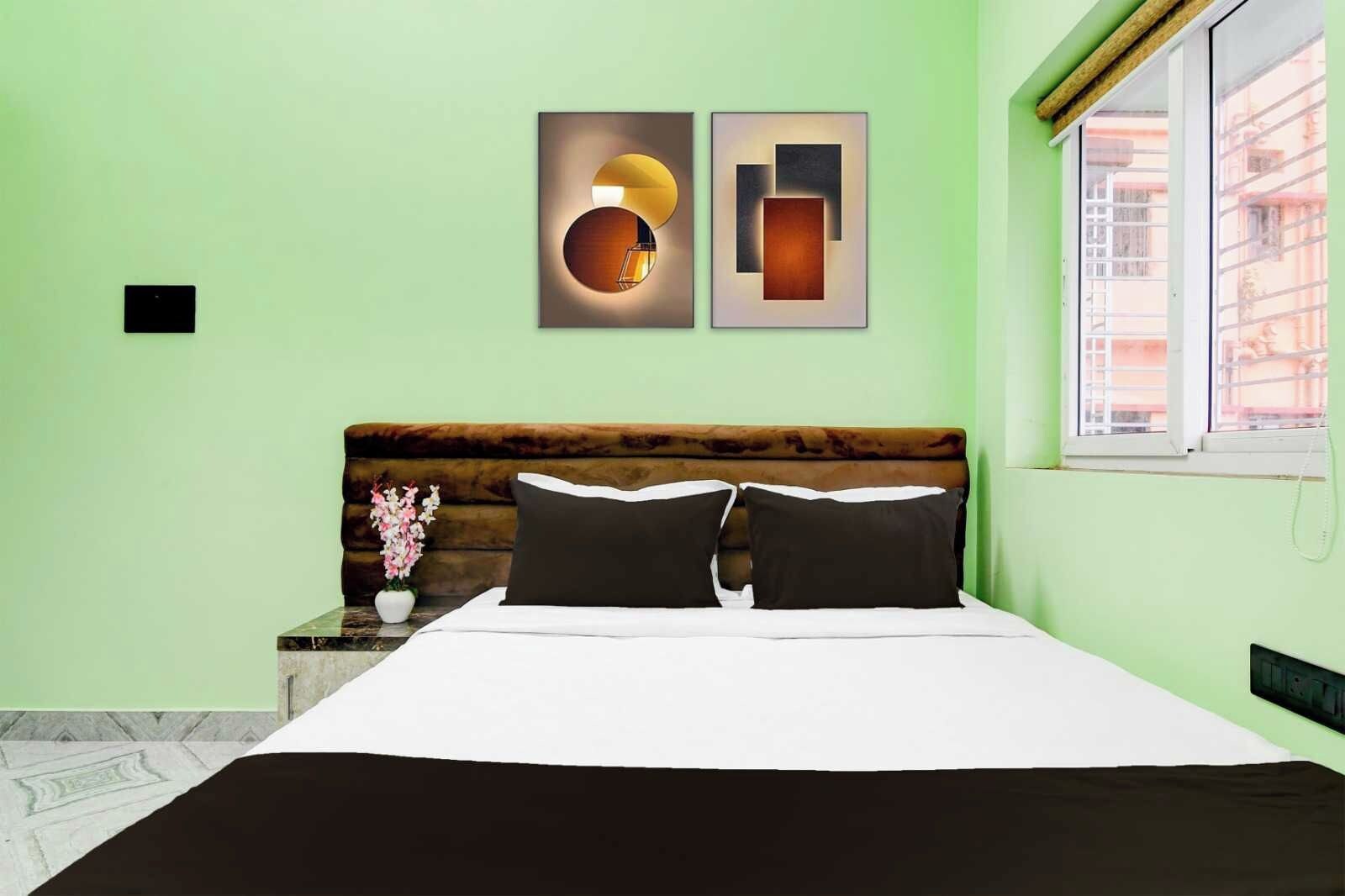 A bedroom with a bed, a nightstand with a vase of flowers, a window with a view, and two wall art pieces.