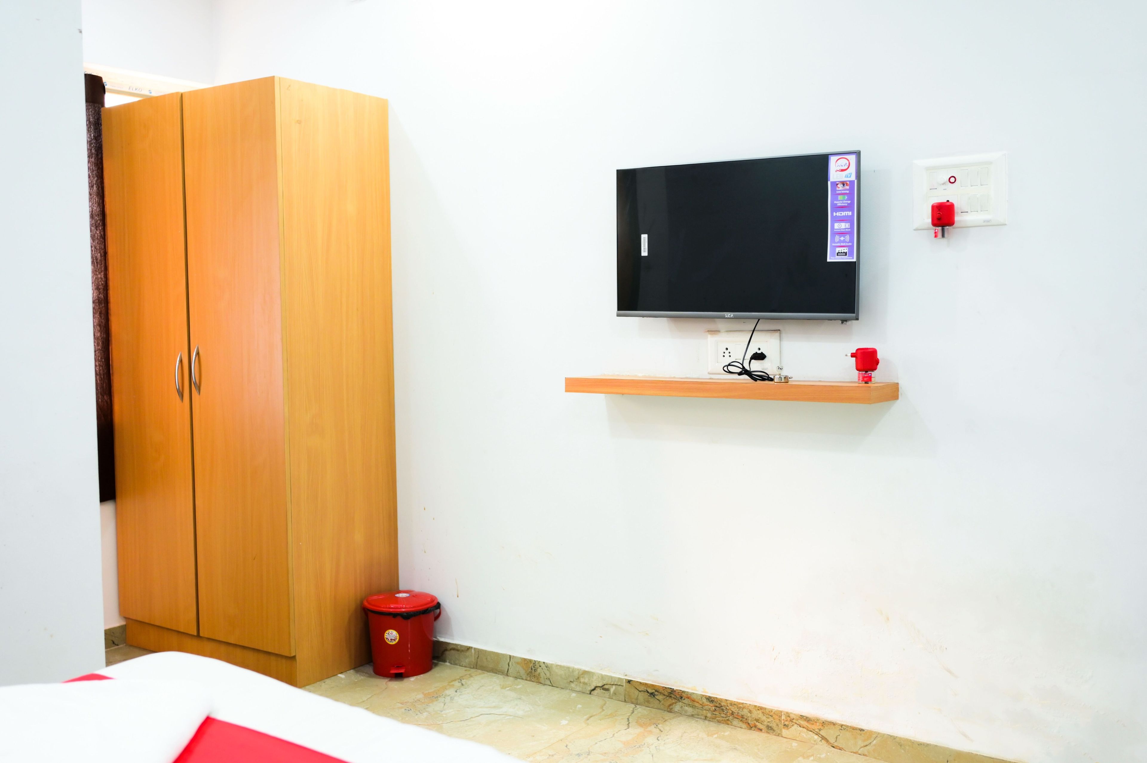 A room with a wooden wardrobe, a television mounted on the wall, a red bucket, and a power outlet with a red socket.