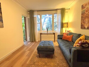 A living room with a sofa, ottoman, and a patterned rug. There is a window with curtains, a lamp, and a painting on the wall. - Chic Ground Floor 2BDR Apartment in Ashfield (Ashfield)