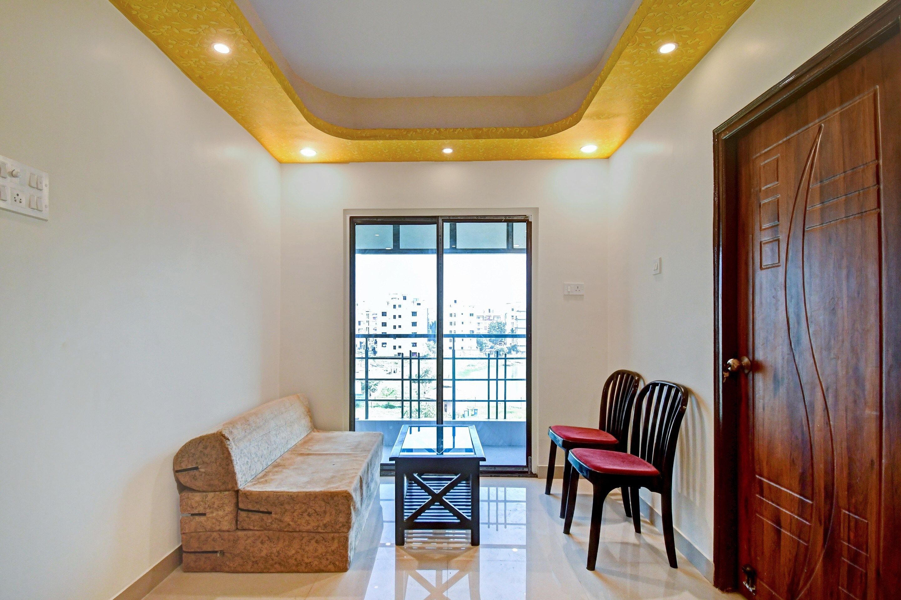Una habitación con sofá, dos sillas, una mesa, una puerta de madera y una ventana con vista a edificios.