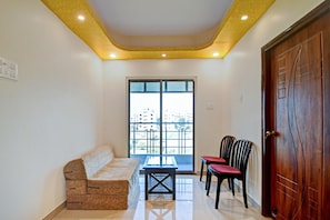 A room with a sofa, two chairs, a table, a wooden door, and a window with a view of buildings. - Hotel O Comfort N Cozy (Kolkata)