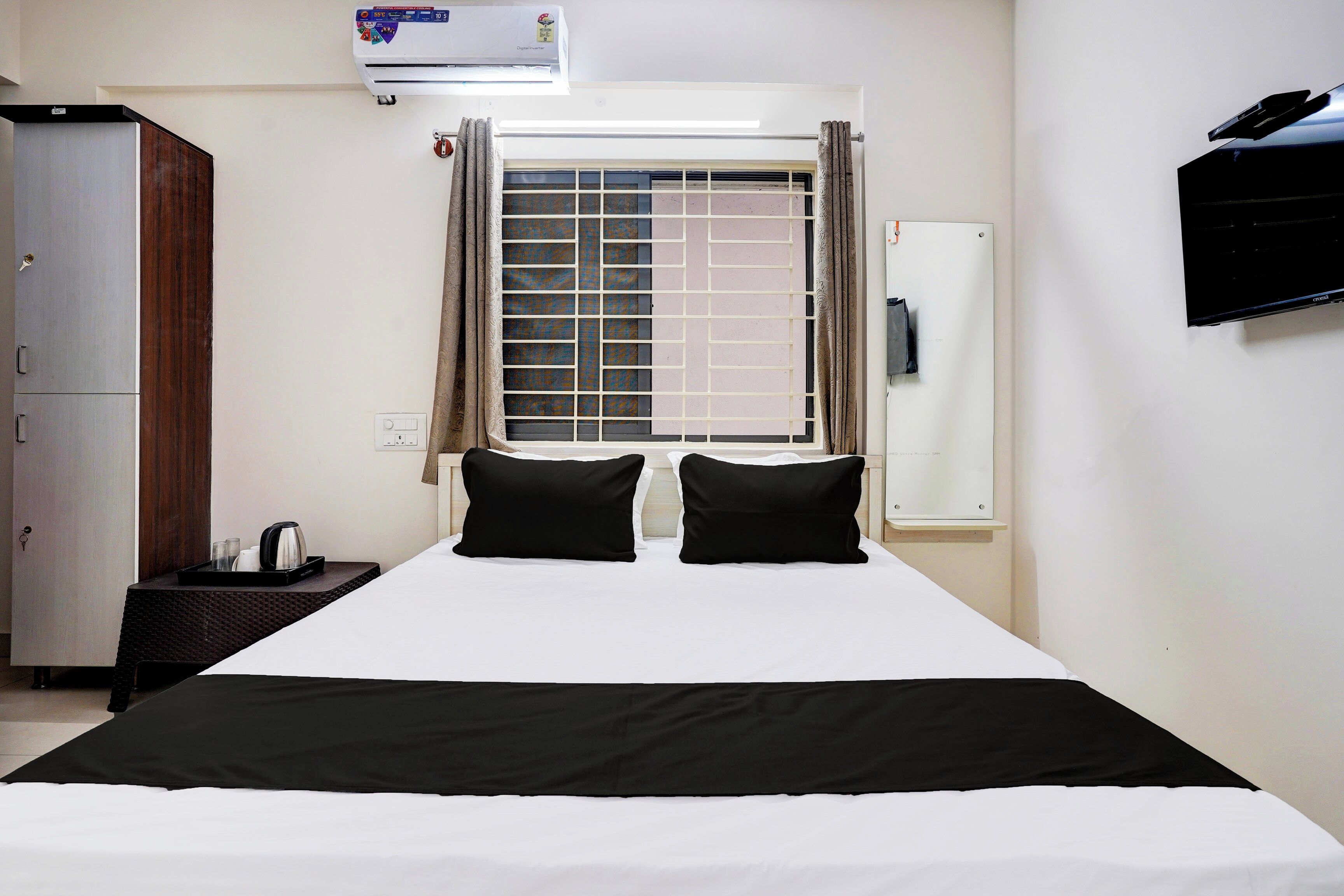 A hotel room with a bed, a wardrobe, a window with curtains, a wall-mounted TV, and an air conditioning unit.