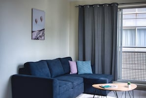 Ein modernes Wohnzimmer mit einem dunkelblauen Sofa, einem kleinen runden Tisch mit einer Zeitschrift und einem Fenster mit Blick auf ein Gebäude.