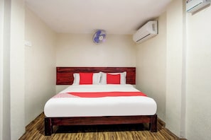 Habitación de hotel con cabecera de madera, cama con ropa de cama blanca y roja, ventilador de pie y unidad de aire acondicionado.