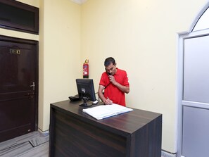 Un hombre con camisa roja de pie frente a un escritorio, con una computadora y un extintor en la pared.