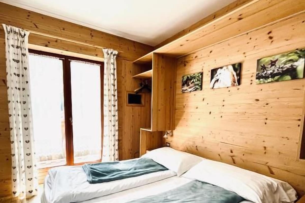 Una habitación con dos camas, paredes de madera, un ventanal con cortinas y un pequeño estante con fotografías.