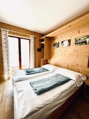 Una habitación con dos camas, paredes de madera, un ventanal con cortinas y un pequeño estante con fotografías.