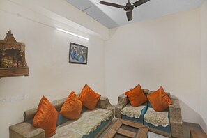 A living room with two sofas, orange cushions, a ceiling fan, a wall-mounted light, and a framed picture. - Hotel O Golden (Noida)
